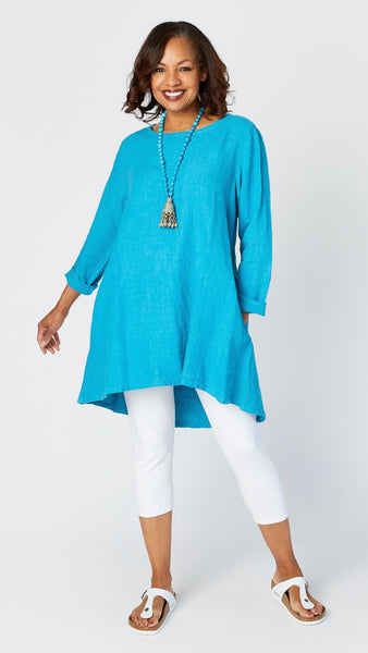 Model wearing summer turquoise high-low linen tunic with long sleeves, turquoise Jhumka necklace, white capri leggings, and sandals. 