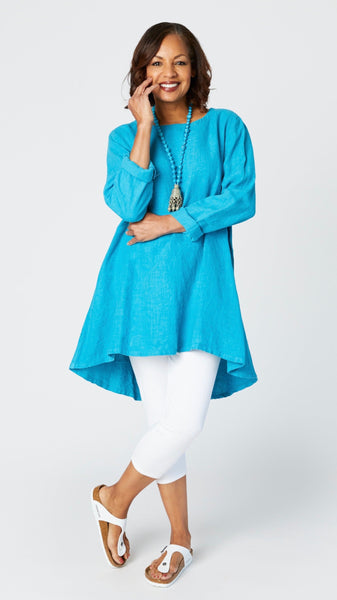 Model wearing summer turquoise high-low linen tunic with long sleeves, turquoise Jhumka necklace, white capri leggings, and sandals.
