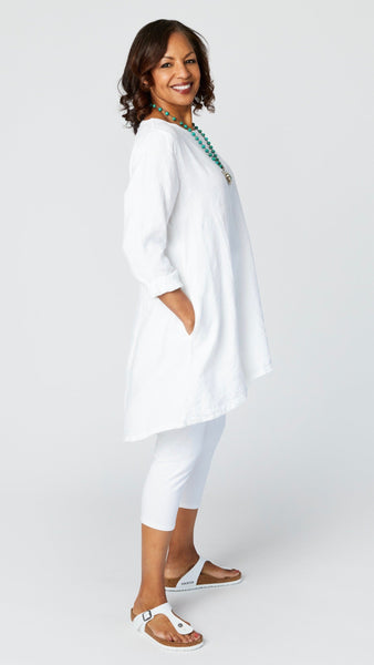 Model wearing white high-low linen tunic with long sleeves, white capri leggings, turquoise pendant necklace, and white sandals.