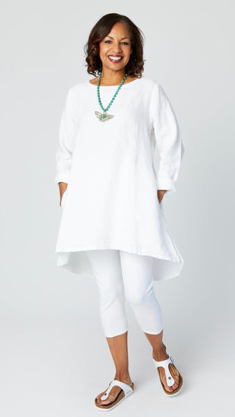 Model wearing white high-low linen tunic with long sleeves, white capri leggings, turquoise pendant necklace, and white sandals.