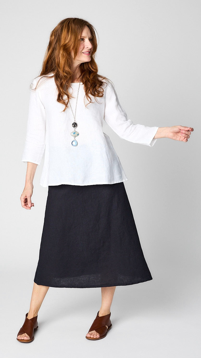 Model wearing white linen 3/4 sleeve shirt with A-line silhouette, ink A-Line Linen skirt, triple stone pendant necklace, and leather sandals. 