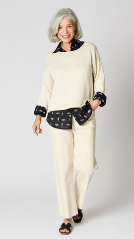 Model wearing cream boxy cotton sweater with drop shoulder and slightly cropped hemline, layered over black button-up shirt, and cropped ecru wideleg jeans. 