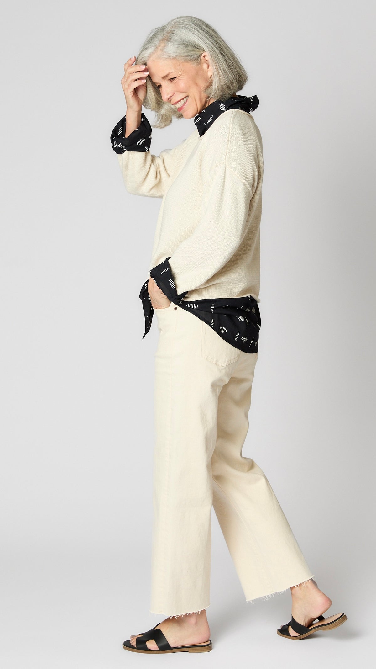 Model wearing cream boxy cotton sweater with drop shoulder and slightly cropped hemline, layered over black button-up shirt, and cropped ecru wideleg jeans.