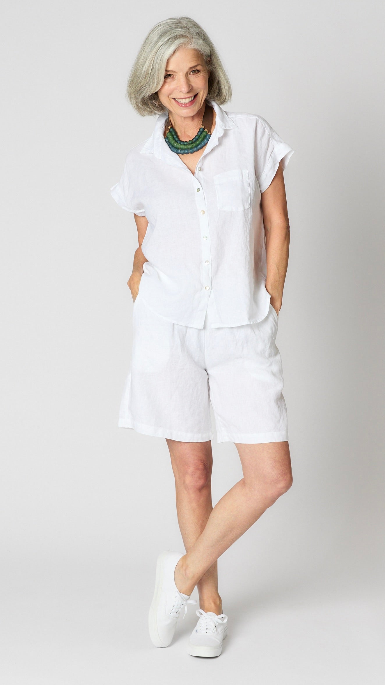 Model wearing white linen capsleeve  camp shirt with mother of pearl buttons, and knee length white linen shorts.  