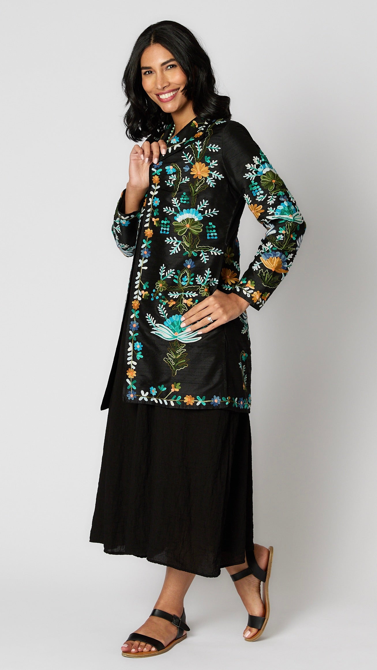 Model wearing black silk duster embroidered with bright blue, yellow and green tones over black rayon romper and black leather sandals.