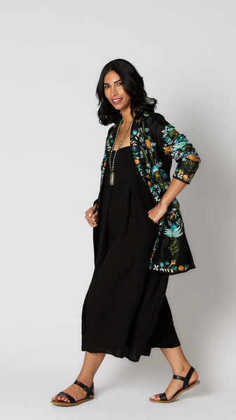 Model wearing black rayon jumpsuit with button shoulder straps, pleat details, and wide cropped leg with embroidered duster, and  black leather boots.