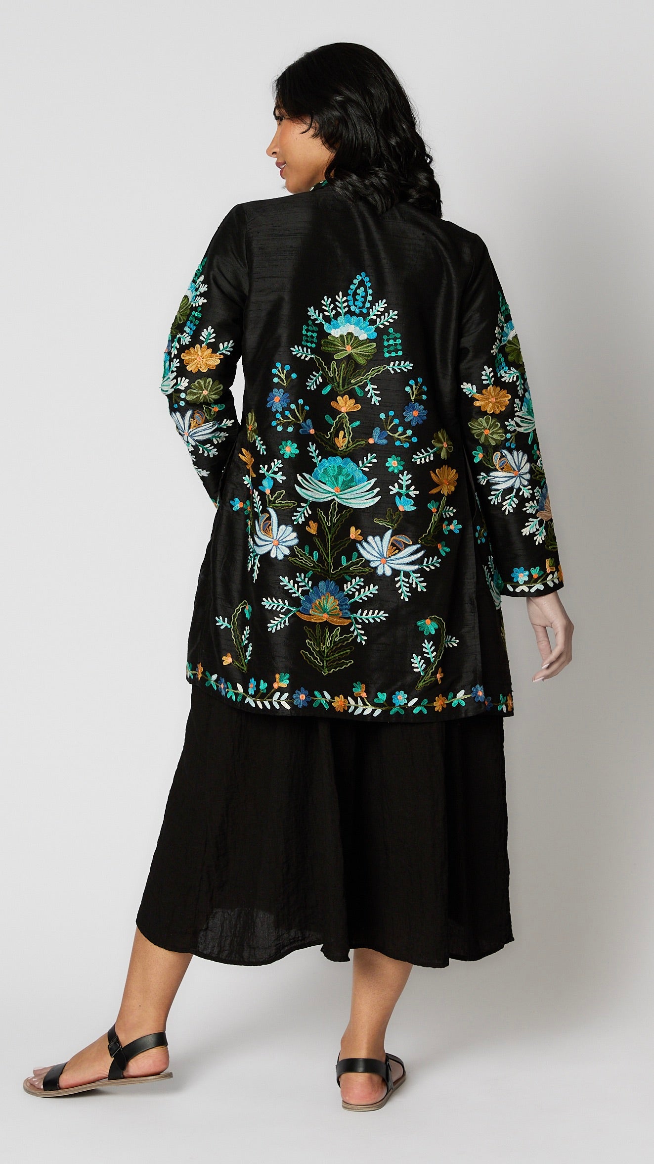 Model wearing black silk duster embroidered with bright blue, yellow and green tones over black rayon romper and black leather sandals.