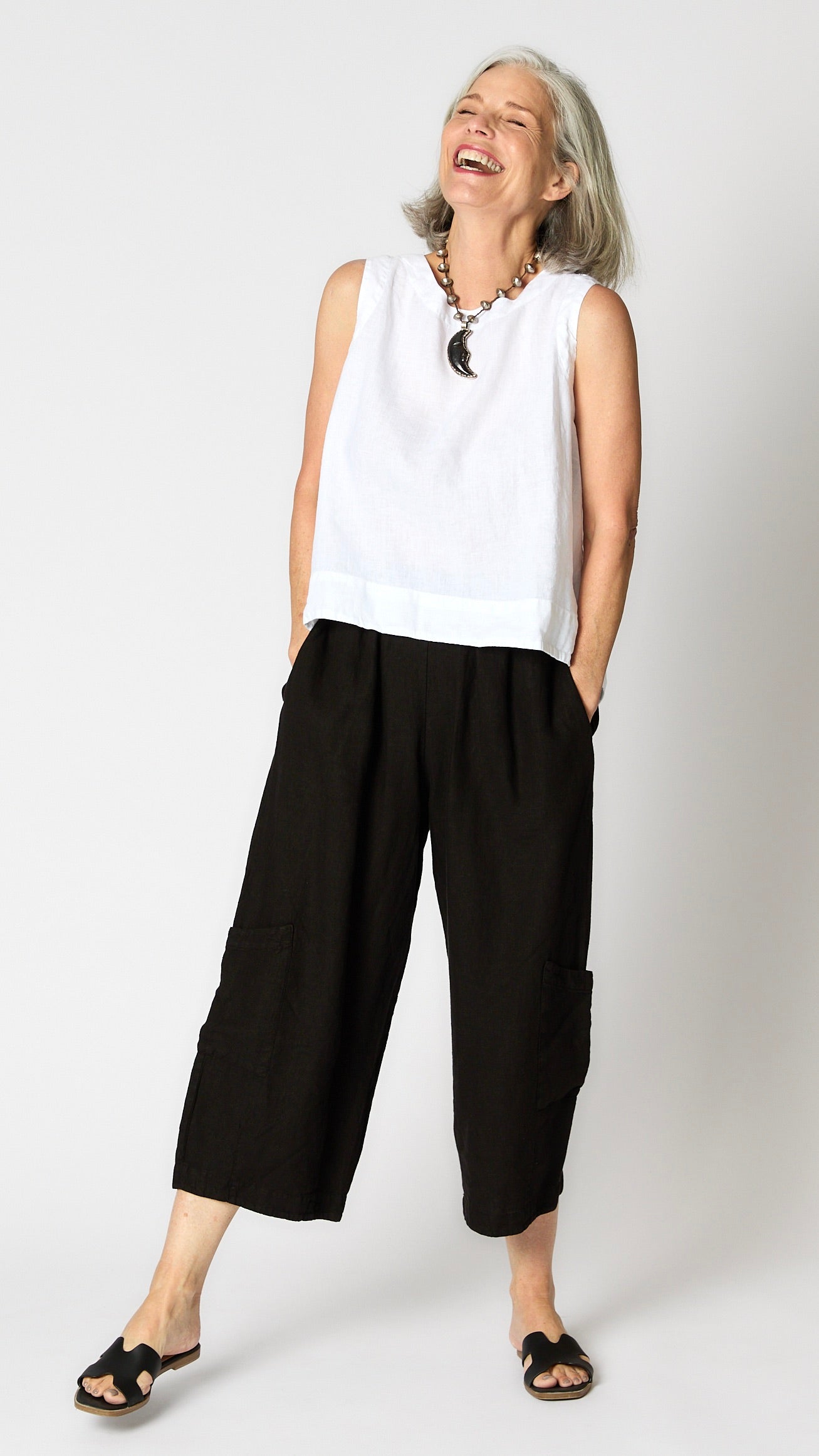 Model wearing white linen hi-lo tank top with dyed bone moon pendant necklace, black cropped linen pants with ankle darts and patch pockets, and black sandals. 