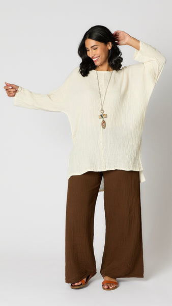 Model wearing chocolate crinkle cotton palazzo pants, cream oversized hi-lo double cotton tunic, and brown leather sandals. 