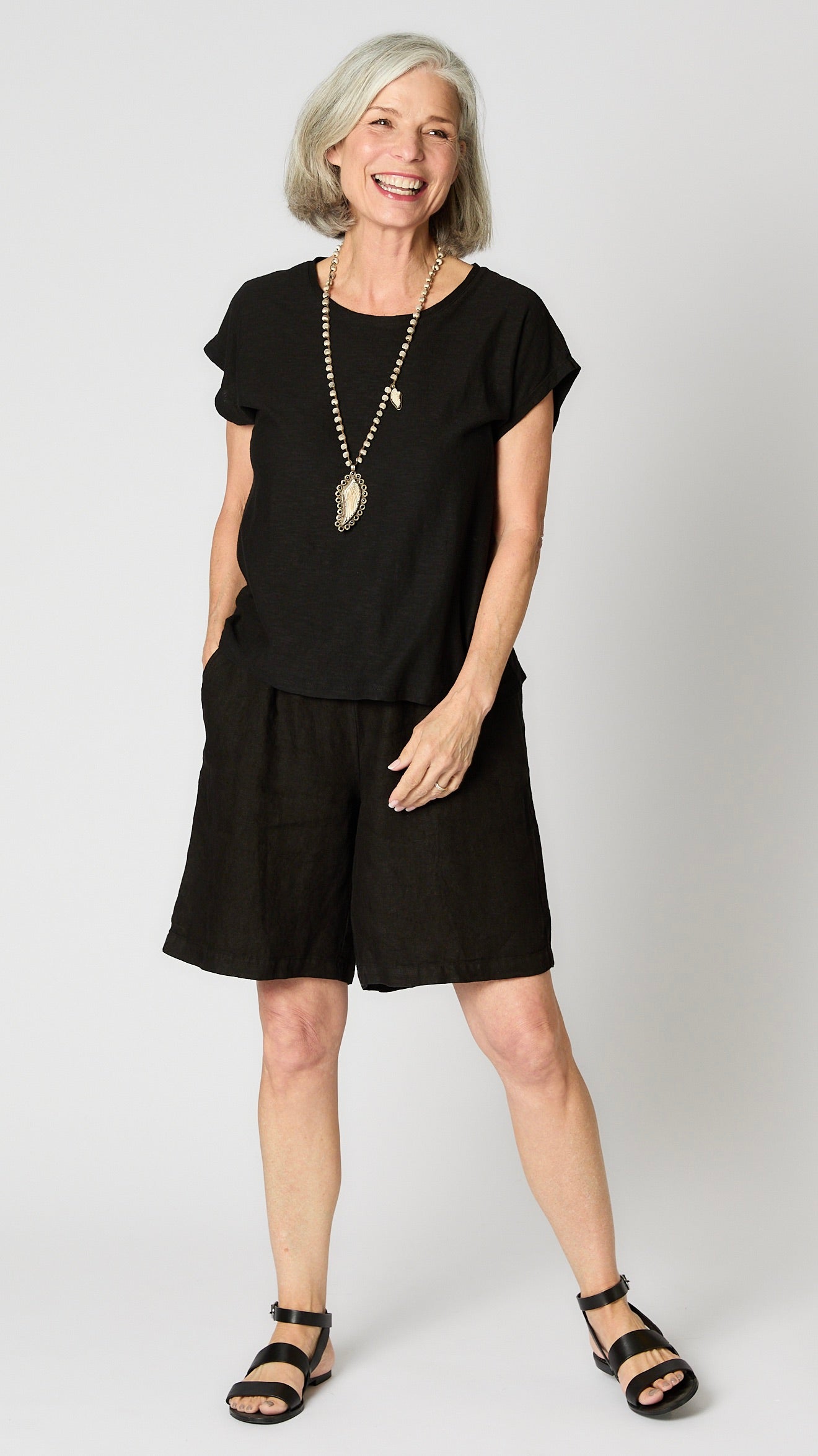 Model wearing black linen blend cropped sleeve  boxy shirt with carved bone pendant necklace and  knee-length black linen shorts.  