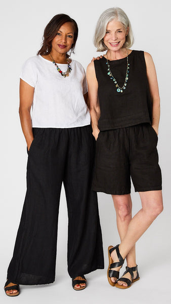 Model wearing colorful charm necklace, white shortsleeve hi-lo top, black full linen pant, black sandals posing next to model in black tank, black knee-length shorts, and black sandals. 