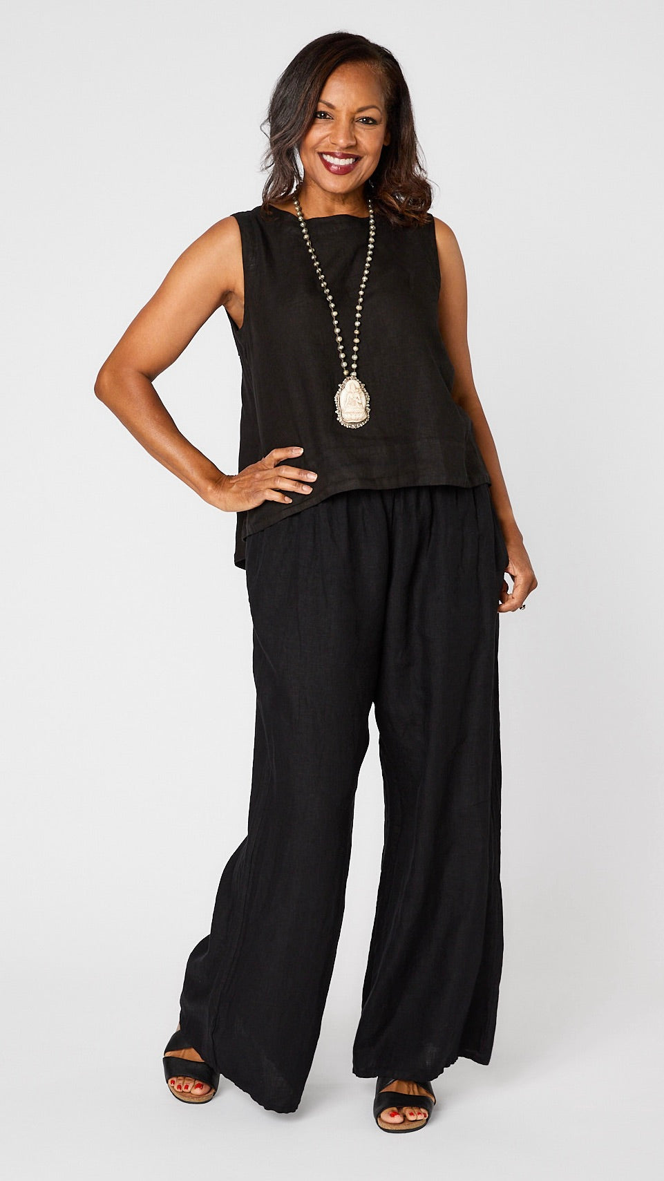 Model wearing black hi-lo linen tank, bone Buddha pendant necklace, and black linen wideleg long pants with black sandals. 