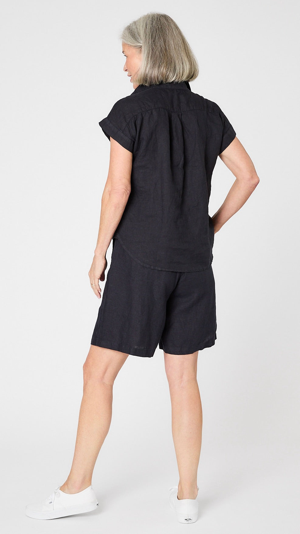 Rearview of model wearing ink linen cap sleeve camp shirt with mother of pearl buttons, and knee length ink linen shorts.  