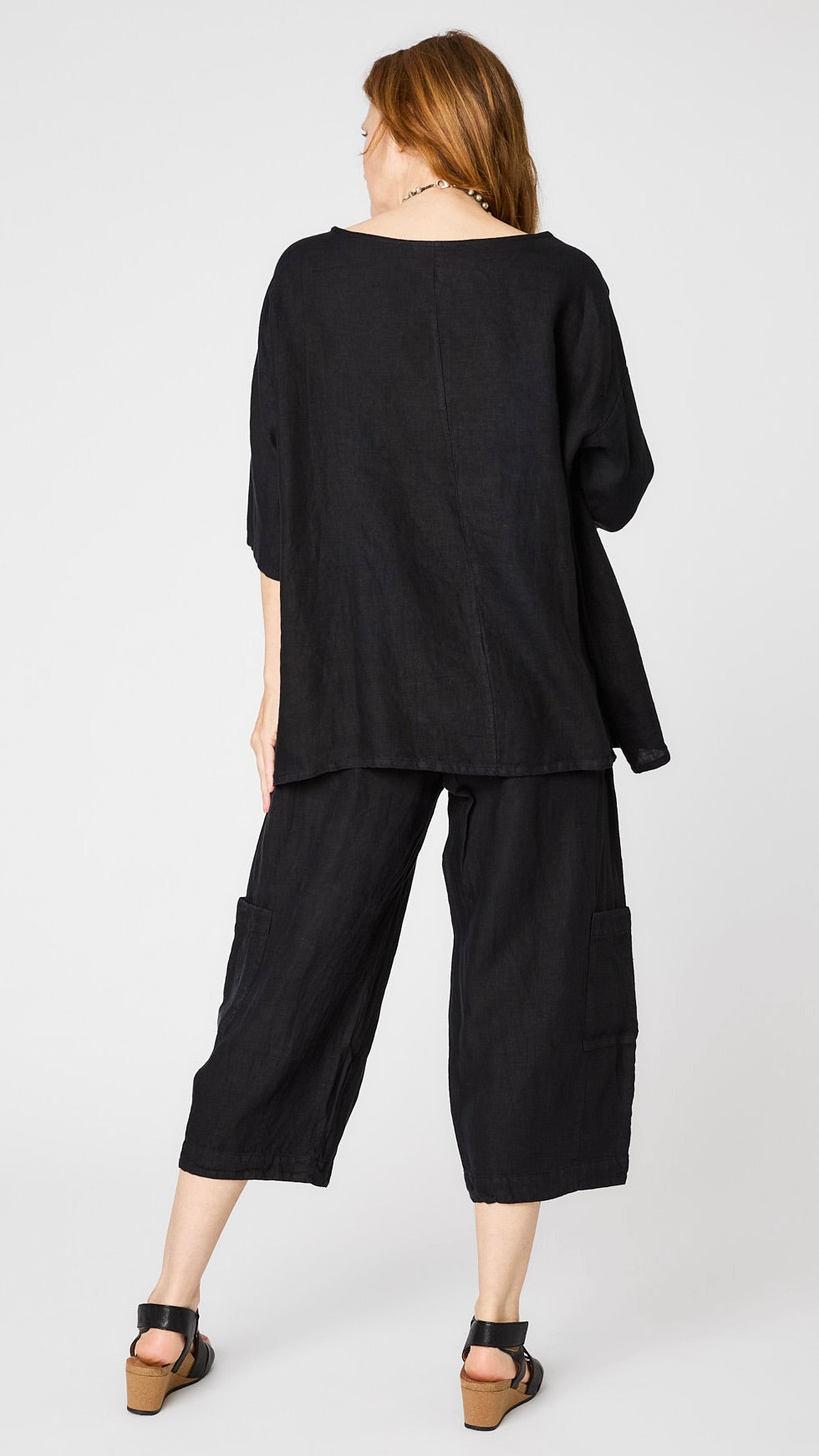 Rearview of model wearing black linen boatneck top with 3/4 sleeves and generous hemline, black cropped linen pants with patch pockets, and black sandals. 