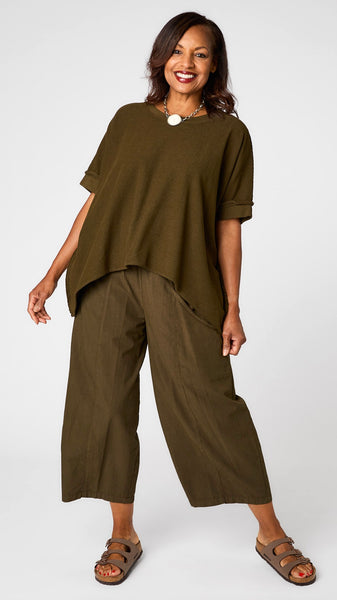 Model wearing African Olive short sleeve pullover vest with oversized drop shoulder, crew neck, and curved hi-lo hemline over African olive one-pocket pant, and brown sandals. 
