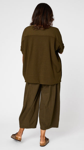 Rearview of model wearing African Olive short sleeve pullover vest with oversized drop shoulder, crew neck, and curved hi-lo hemline over African olive one-pocket pant, and brown sandals. 