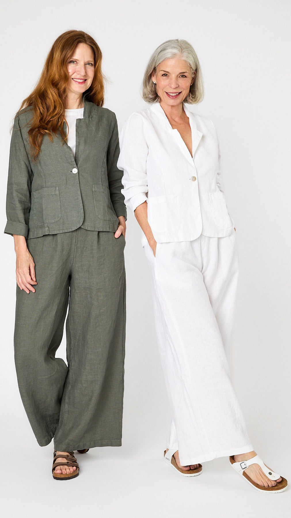 Two models pose together - one wears the linen blazer and wide leg linen pant set in white, and the other wears the set in  "clay" green-gray. 