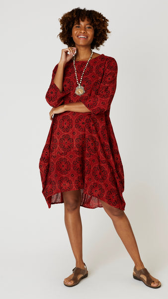 Model wearing "rust medallion" block print tunic dress with 3/4 sleeves, high-low hemline and bubble silhouette, with bone bead and pendant necklace, and brown leather sandals.