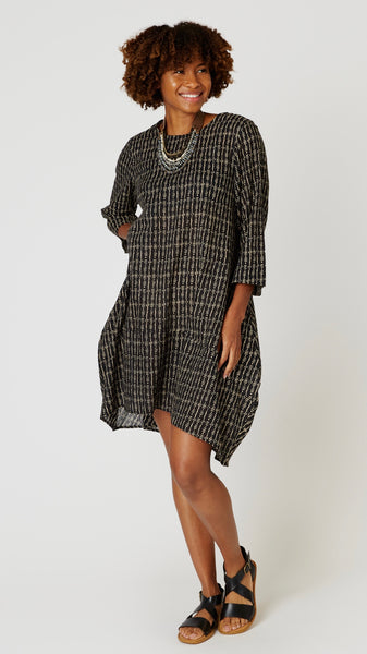 Model wearing "tribal black" block print tunic dress with 3/4 sleeves, high-low hemline and bubble silhouette, with beaded leather necklace, and black leather sandals.