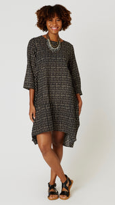 Model wearing "tribal black" block print tunic dress with 3/4 sleeves, high-low hemline and bubble silhouette, with beaded leather necklace, and black leather sandals.