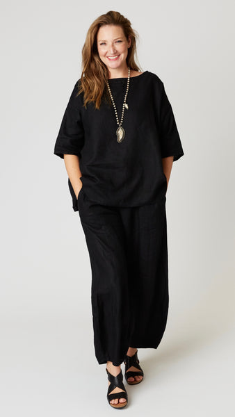 Model wearing black linen boatneck top with 3/4 sleeves and generous hemline, with black linen lantern pants, and black leather sandals.