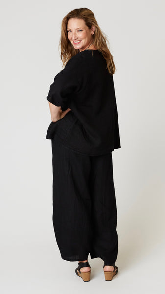 Model wearing black linen boatneck top with 3/4 sleeves and generous hemline, with black linen lantern pants, and black leather sandals.