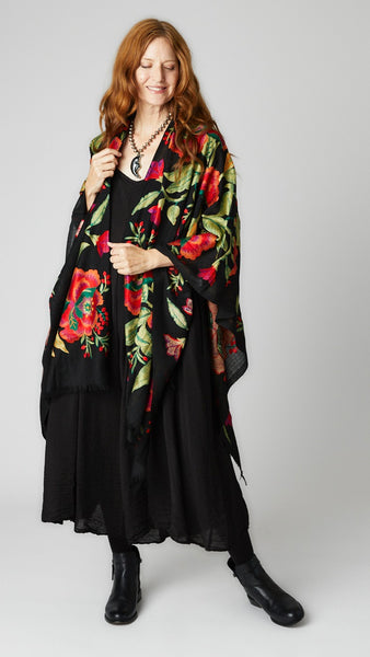 Model wearing black embroidered wool ruana with vibrant red and green florals, black rayon romper, and black leather ankle boots.