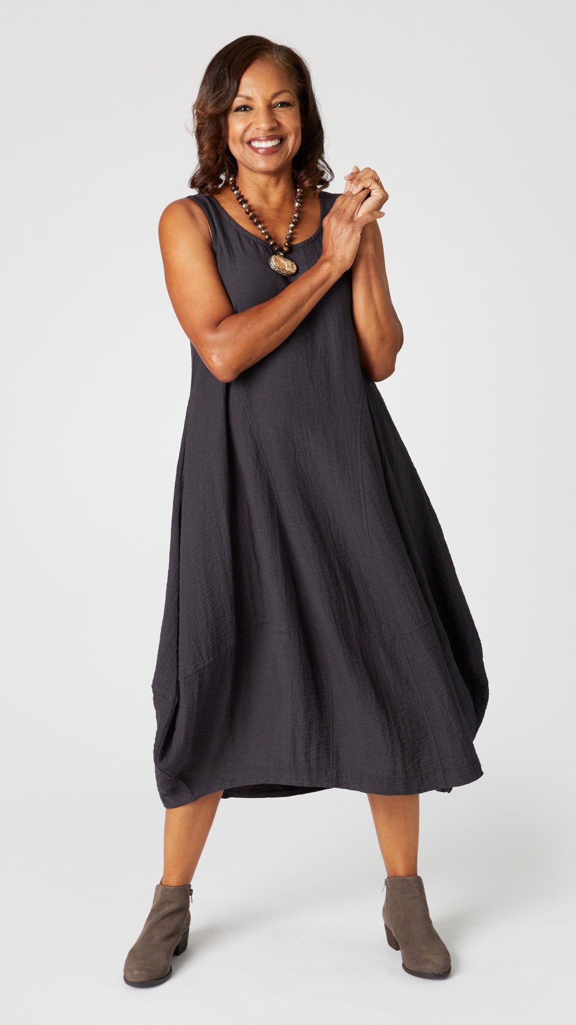Model wearing "black pearl" sleeveless rayon dress with tulip hemline and round neck, beaded necklace with bone pendant, and gray suede boots. 