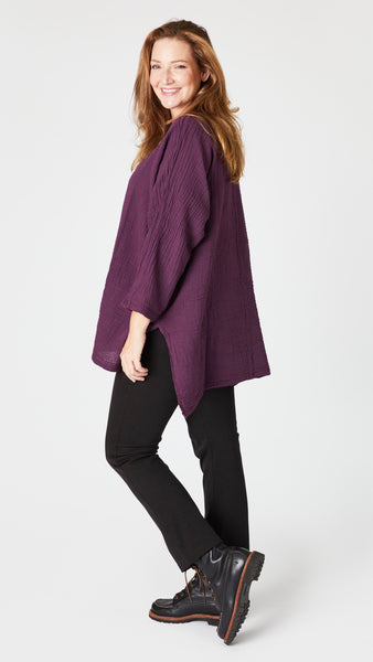 Model wearing eggplant 3/4 sleeve, split hem, flowy high-low tunic in double cotton, with black straight leg jeans, and black lace-up leather boots.