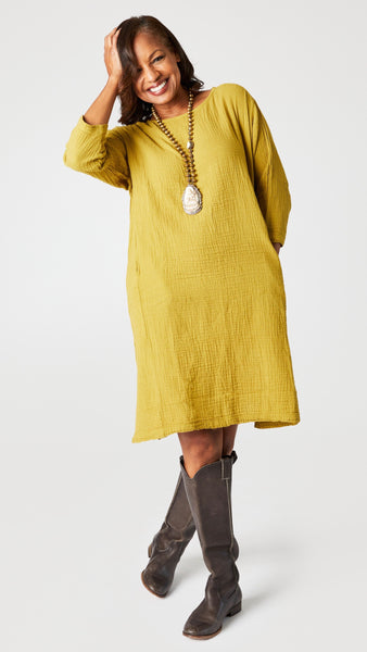 Model wearing ochre crop-sleeve knee length cotton dress with side seam pockets, brass bead and bone pendant necklace, knee high leather boots.