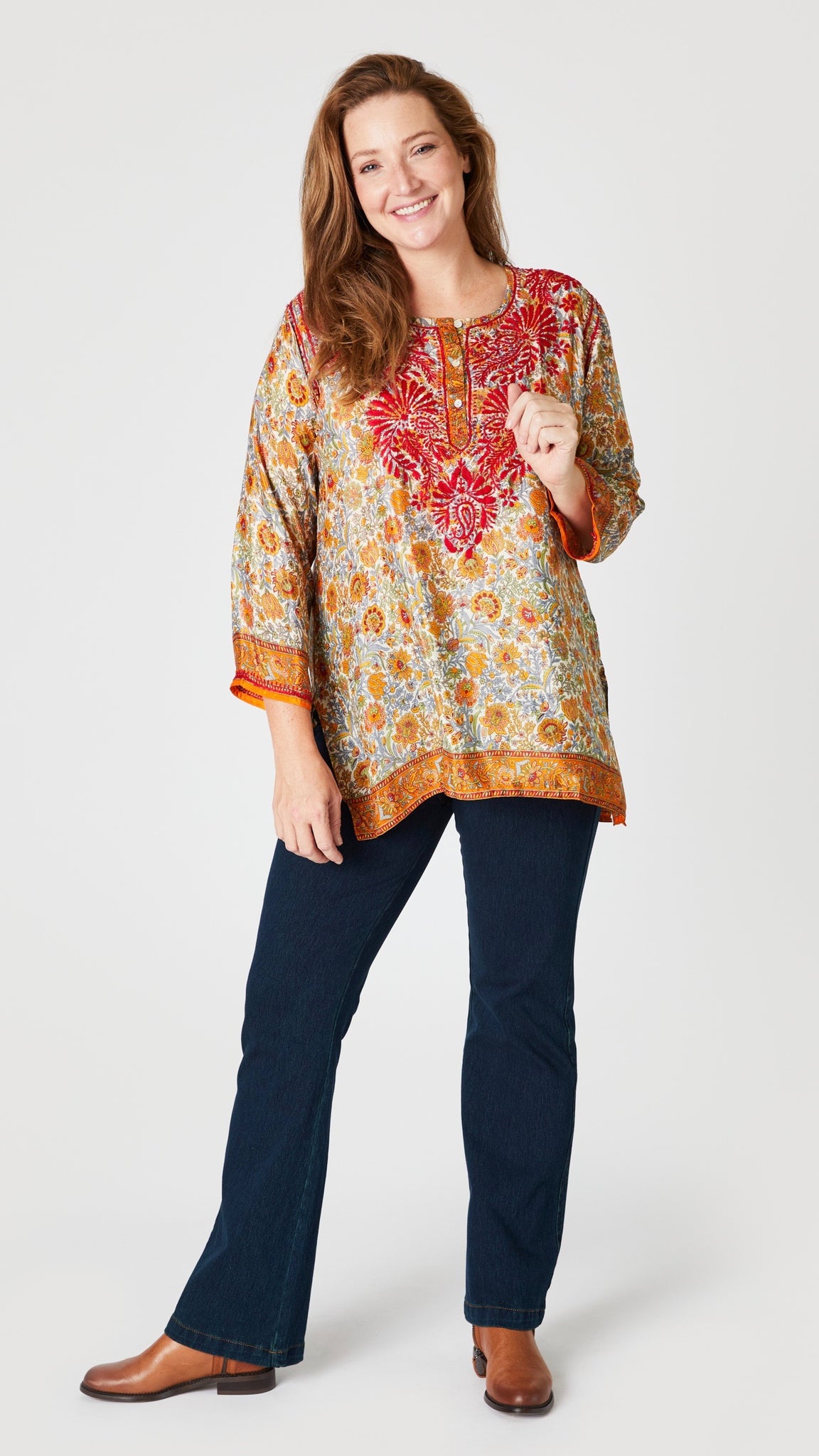 Model wearing amber and red silk embroidered tunic with 3/4 sleeves, indigo bootcut jeans, and tan leather boots.