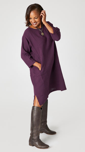 Model wearing eggplant crop-sleeve knee length cotton dress with side seam pockets, coin pendant necklace, knee high leather boots. 