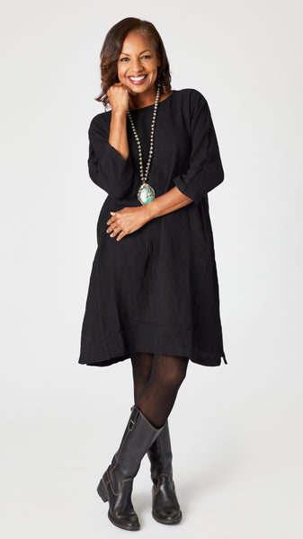 Model wearing black crop-sleeve knee length cotton dress with side seam pockets, turquoise pendant necklace, black sheer tights, knee high leather boots.