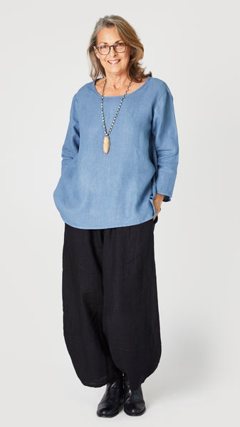 Model wearing denim blue linen A-line top with 3/4 sleeves, black linen lantern pants, and black leather boots.