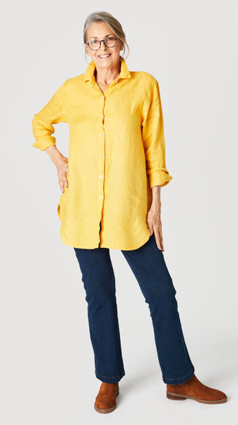 Model wearing sunflower linen button-up shirt with collar and handkerchief hem with indigo straight leg jeans and brown suede boots.