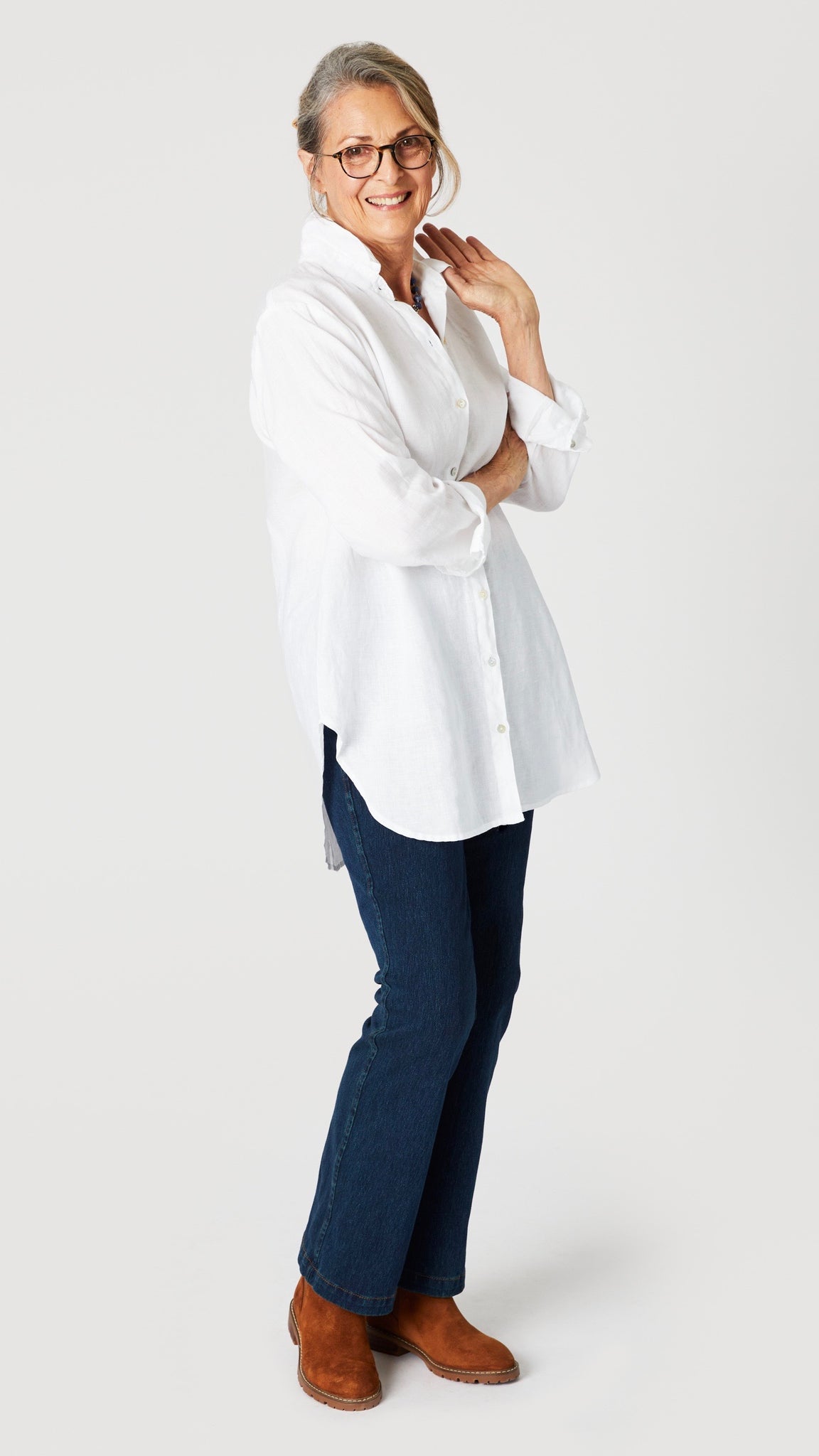 Model wearing white linen button-up shirt with collar and handkerchief hem with indigo bootcut jeans and brown suede boots.