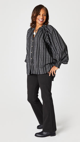 Model wearing black and white vertical striped button up top with gathered collar, elastic cuff sleeves, and flared waist, black bootcut jeans, and black leather boots.