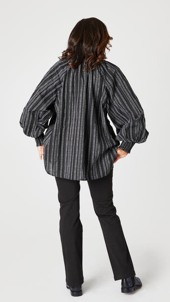 Model wearing black and white vertical striped button up top with gathered collar, elastic cuff sleeves, and flared waist, black bootcut jeans, and black leather boots.