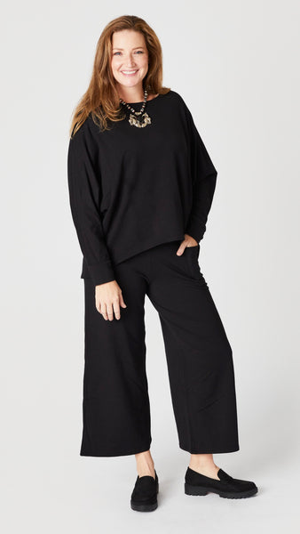Model wearing black boatneck long sleeve top with high low hemline, black cropped palazzo pants, and black lug sole loafers.