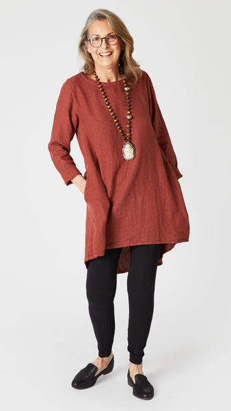 Model wearing cognac high-low linen tunic with long sleeves, brass bead and bone pendant necklace,  black cotton leggings, and black leather loafers.