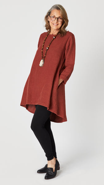 Model wearing cognac high-low linen tunic with long sleeves, brass bead and bone pendant necklace,  black cotton leggings, and black leather loafers.