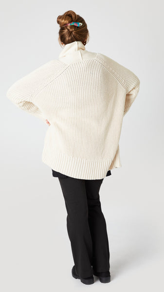 Rear view of model wearing cream cotton handknit oversized cardigan (without buttons) over black jersey tee, black straight leg jean, and black suede loafer. 