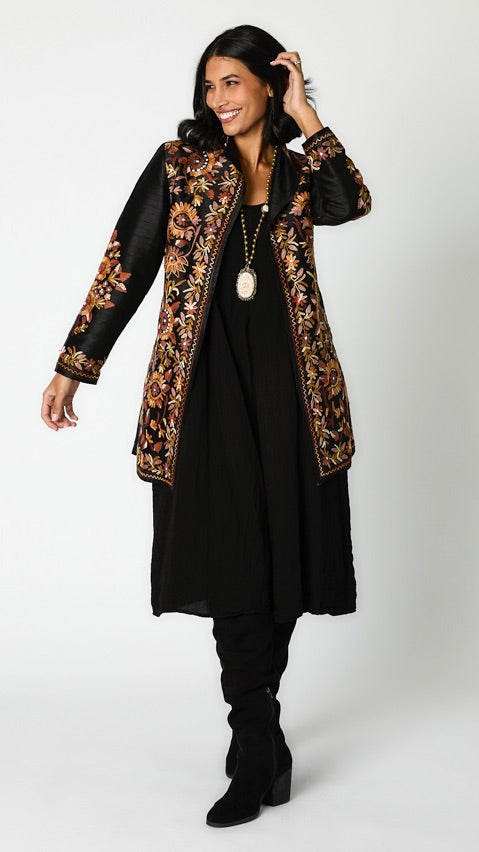 Model wearing black silk jacket with brown toned embroidery over black rayon romper and black suede boots. 