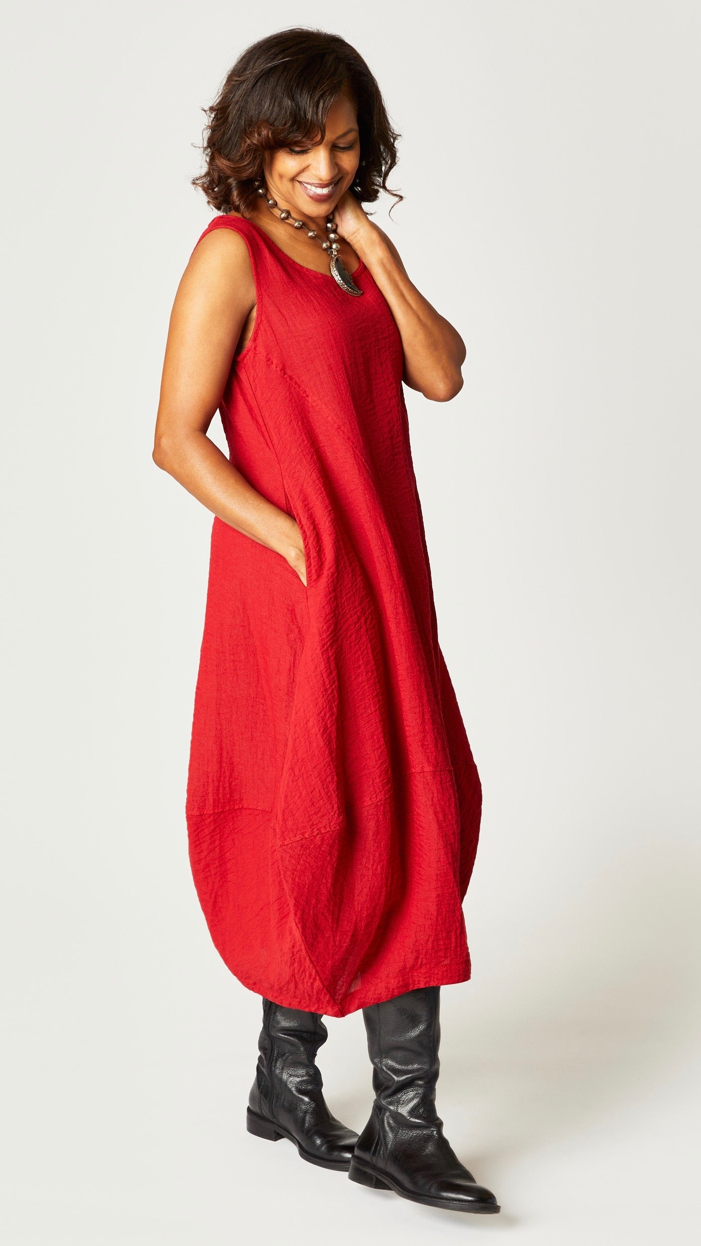 Model wearing "cinnabar" sleeveless rayon dress with tulip hemline and round neck, beaded necklace with dyed bone moon pendant, and black leather boots. 