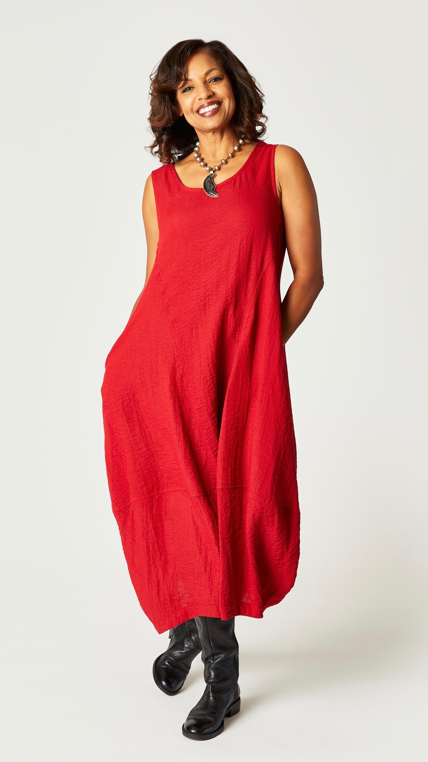 Model wearing "cinnabar" sleeveless rayon dress with tulip hemline and round neck, beaded necklace with dyed bone moon pendant, and black leather boots. 