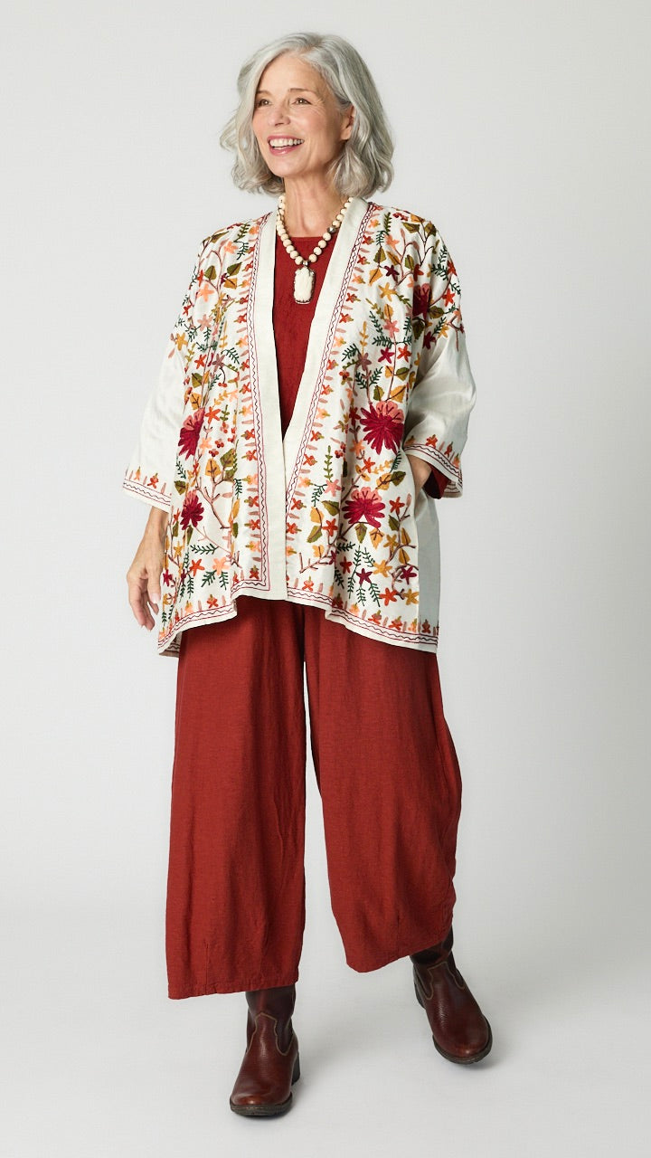 Model wearing white silk kimono style jacket with red, green, yellow floral embroidery, cognac cropped top and cognac cropped dart pants in rayon, carved bone rabbit pendant necklace, and brown leather boots