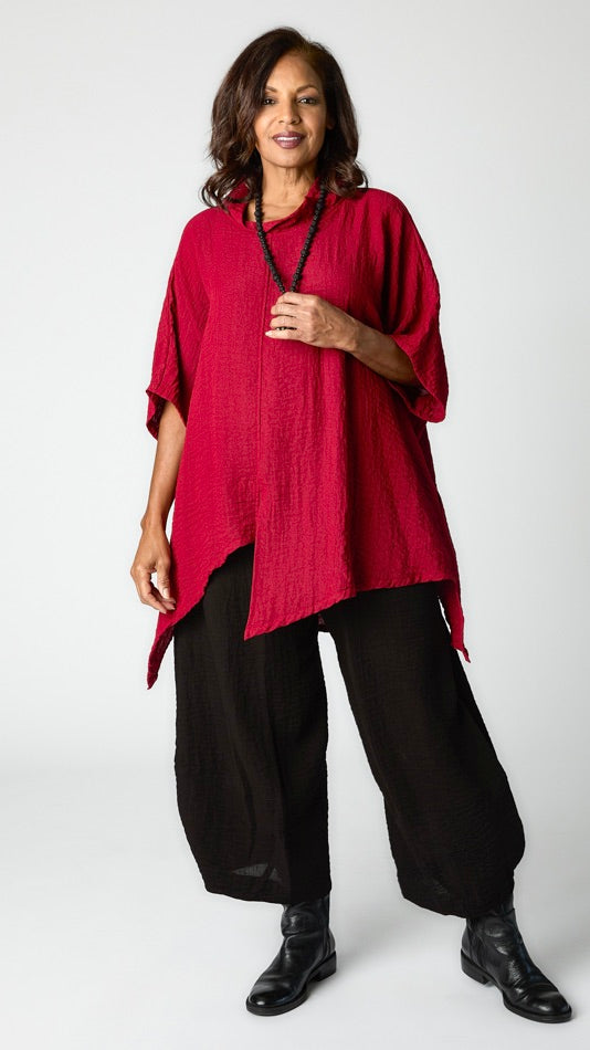 Model wearing black tucked hem cropped pant in travel weight rayon with "masai" asymmetrical cowl neck top in travel rayon, and black leather boots. 