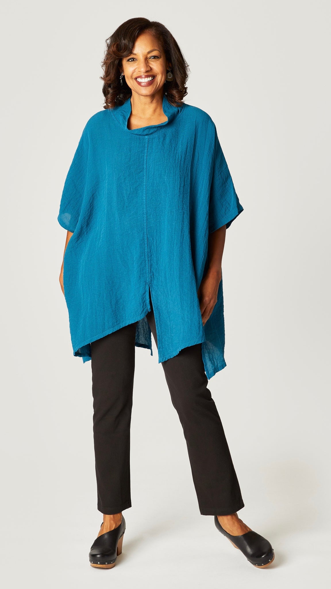 Model wearing teal loose cowl neck tunic with asymmetrical hem and 3/4 sleeves, and black straight leg jeans.