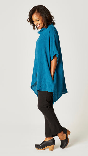 Model wearing teal loose cowl neck tunic with asymmetrical hem and 3/4 sleeves, and black straight leg jeans.