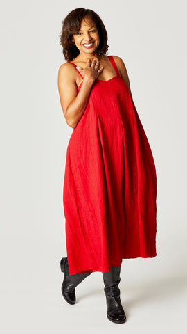 Model wearing red rayon jumpsuit with button shoulder straps, pleat details, and wide cropped leg with black leather boots.